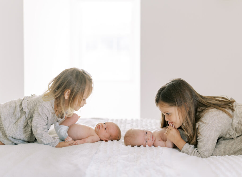 Big sisters looking down at newborn brother and sister twins on a bed