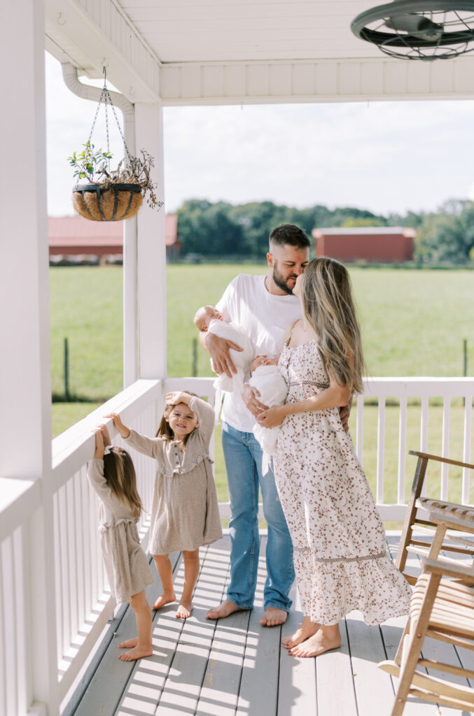 mom and dad kissing while holding newborn twins on front porch with other kids around them
