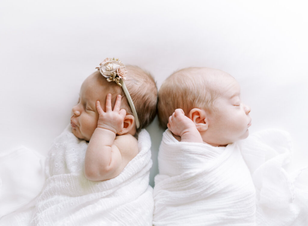 Newborn twins back to back swaddled in all white on white backdrop stretching 