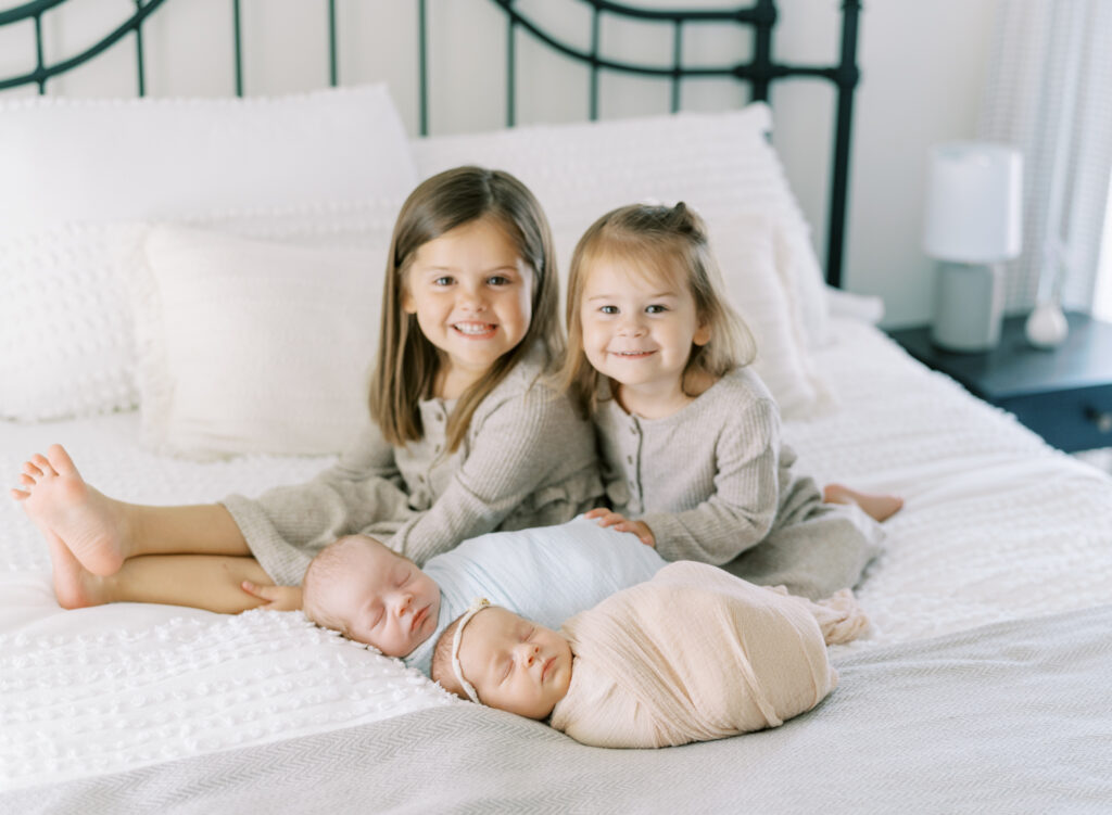 Big sisters and newborn twins brother and sister posed on a bed for a lifestyle newborn photo session. 