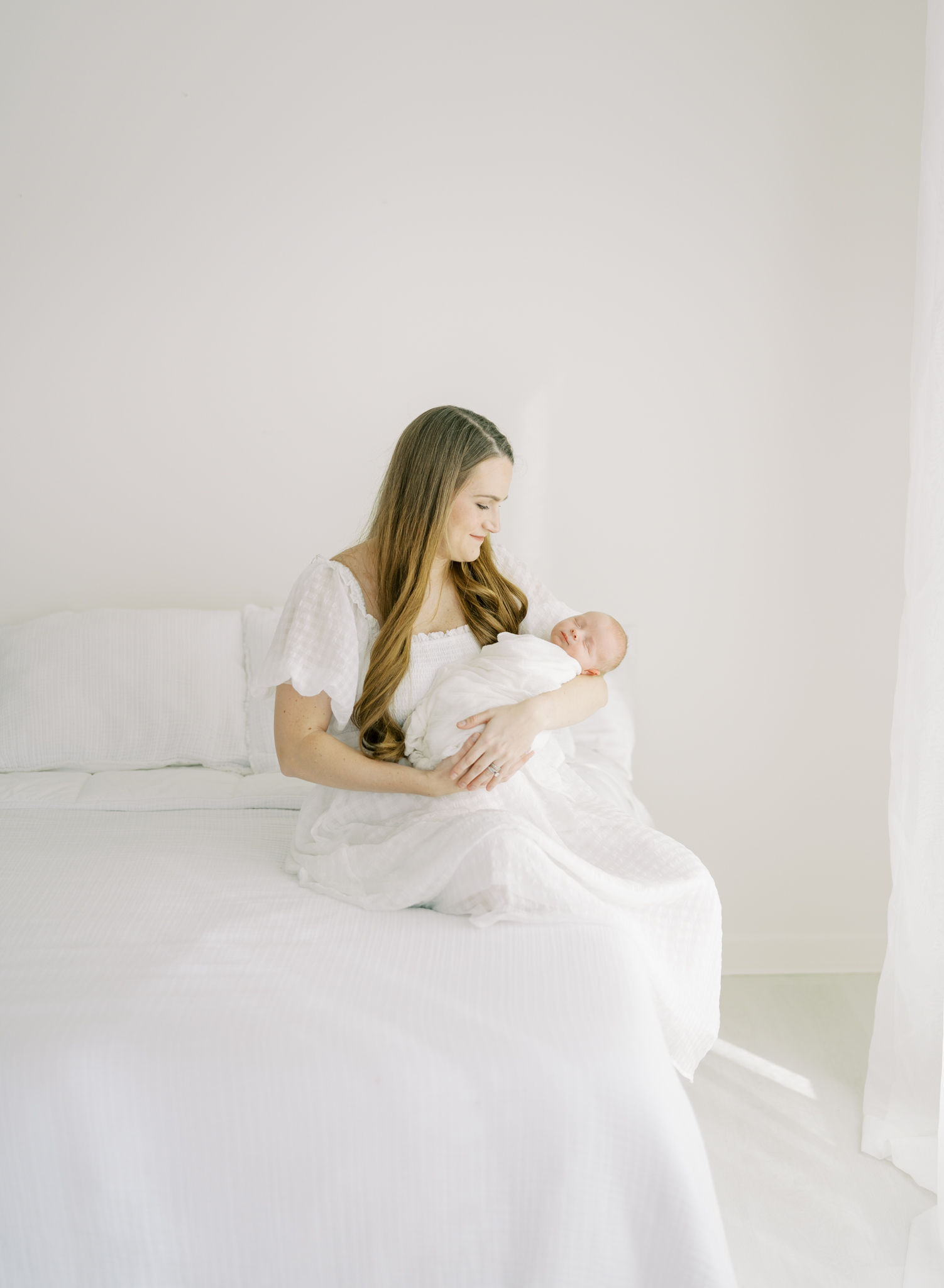 mother sitting on bed holding newborn in sun drenched white room