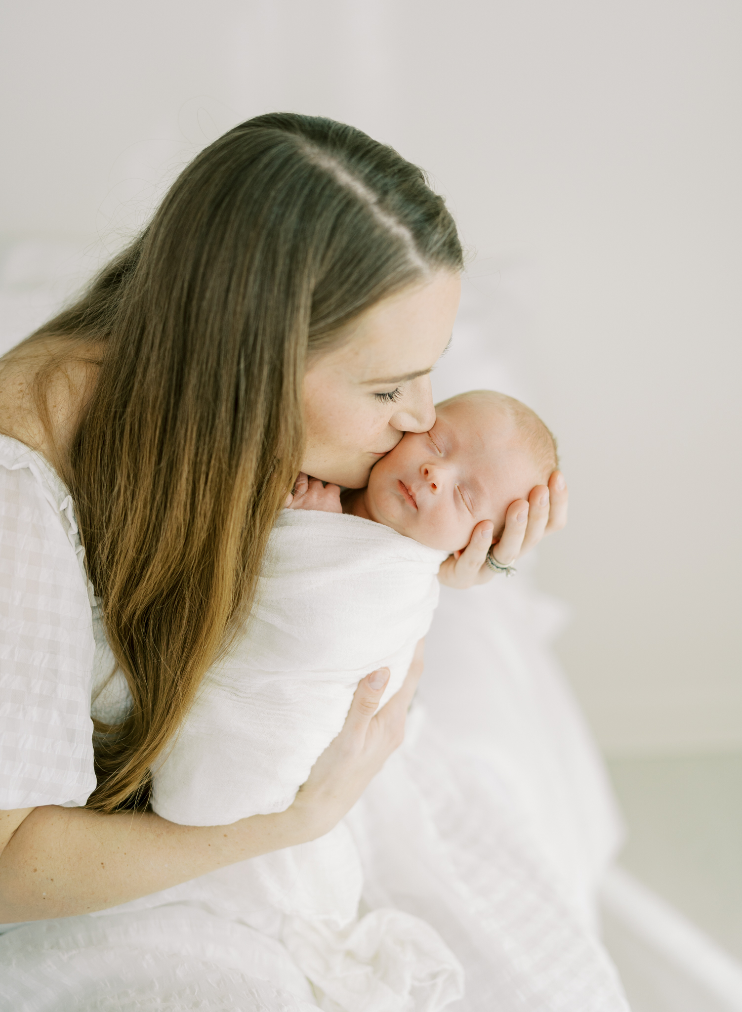 mother kissing newborn son in sun drenched white photography studio