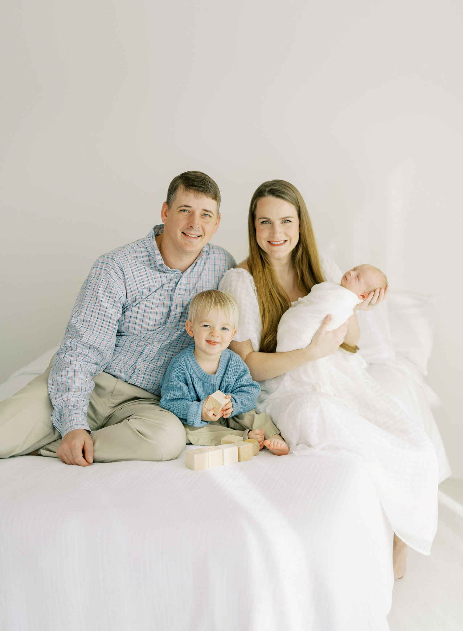 happy family of 4 posing on bed in white studio for newborn photos