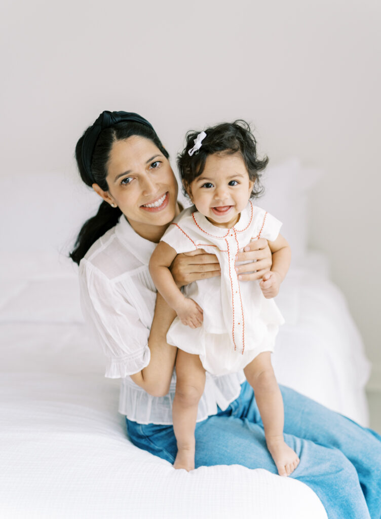 Mother and 6 month old baby girl smiling on a white bed in a clean white studio.