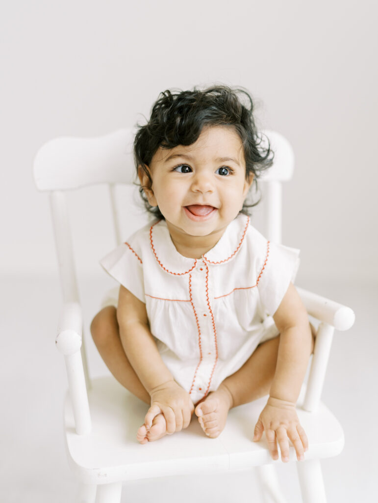 6 month baby girl smiling on a white chair for clean, bright, and natural portrait in Atlanta photography studio.