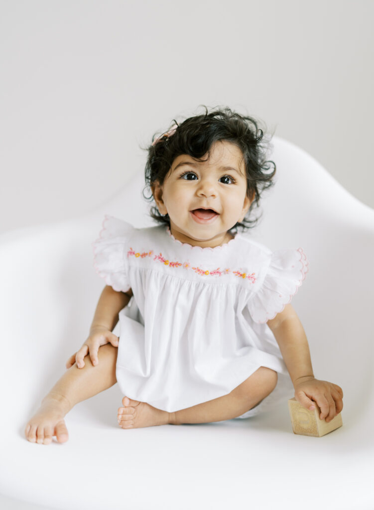 6 month baby girl smiling on a white chair for clean, bright, and natural portrait in Atlanta photography studio.