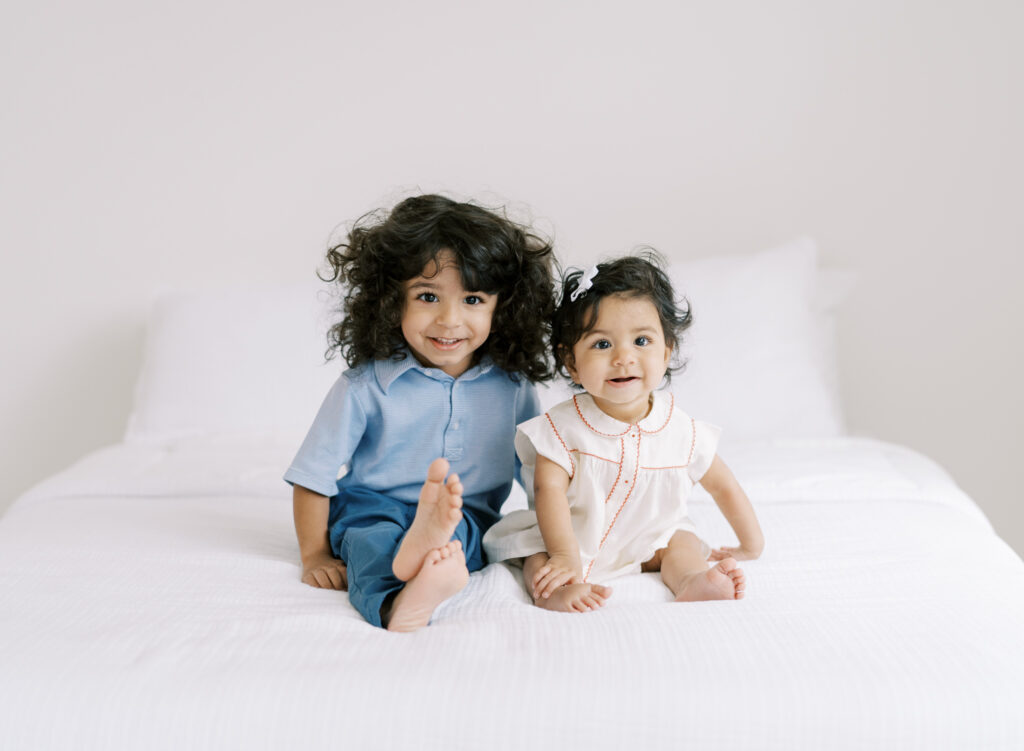 Brother and sister smiling on a white bed in a clean white studio for a natural sibling photo.