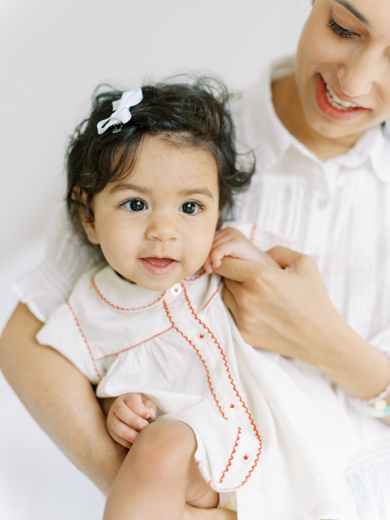 6 month baby girl with soft skin and glistening eyes in her moms arms.