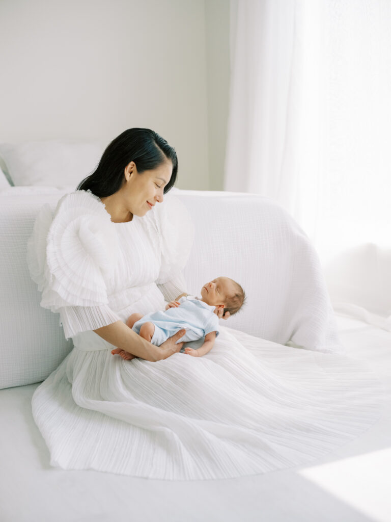 Mom sitting on the floor in an elegant white dress looking down at her newborn son with natural light pouring into the room. 