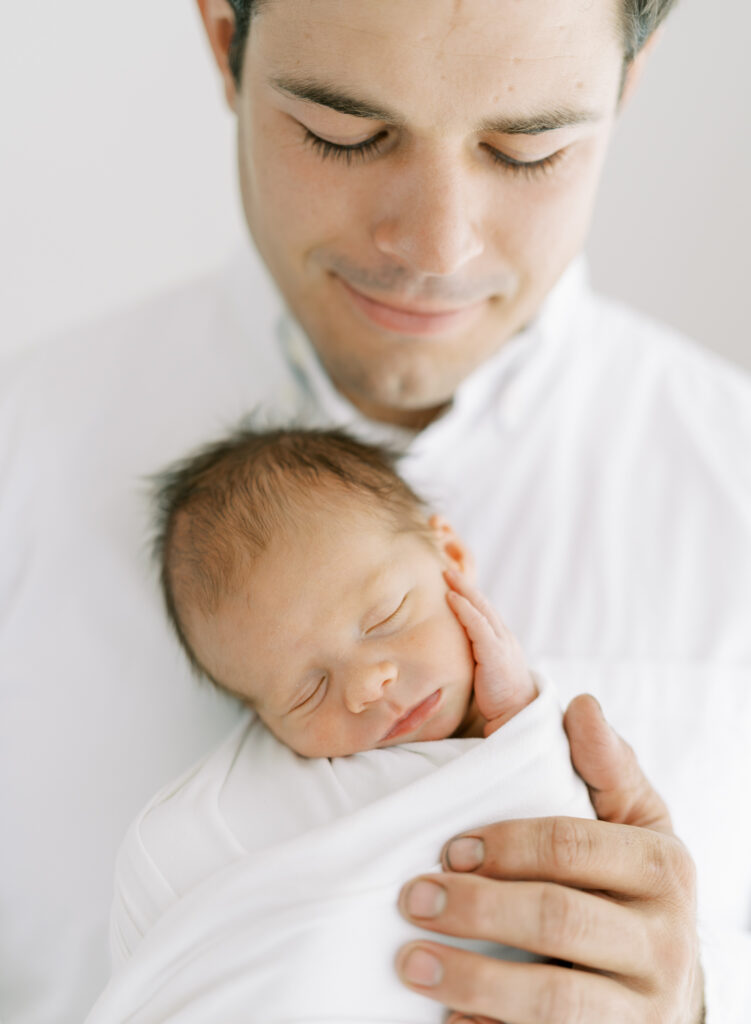 Dad holding tiny newborn son who has his little hand on his cheek in stunning father and son portrait. 