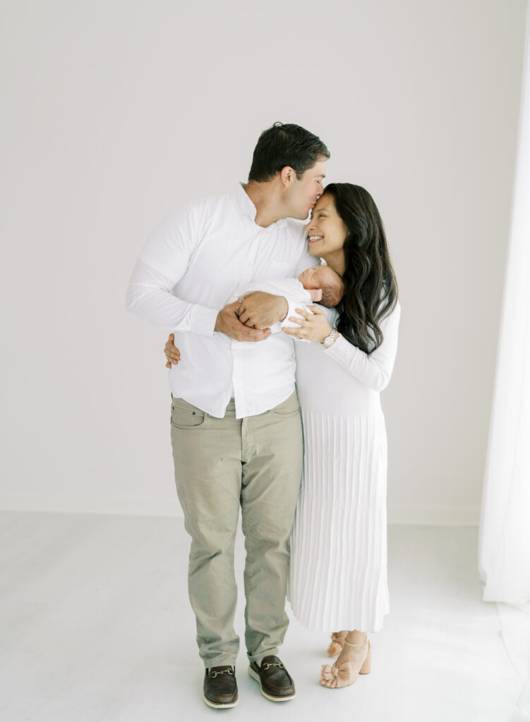 Dad kissing mom while holding newborn son in white photography studio in Cumming, GA. 