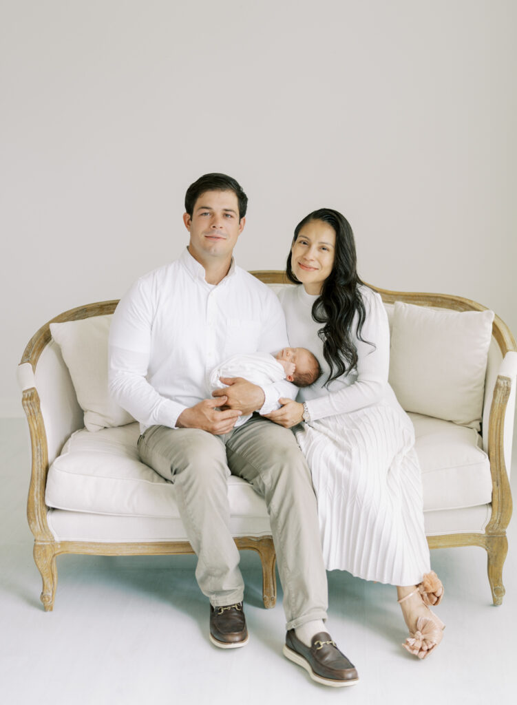 New parents holding newborn son on elegant couch in white photography studio in Cumming GA.