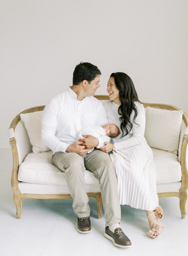 New parents holding newborn son and laughing on elegant couch in white photography studio in Cumming GA.