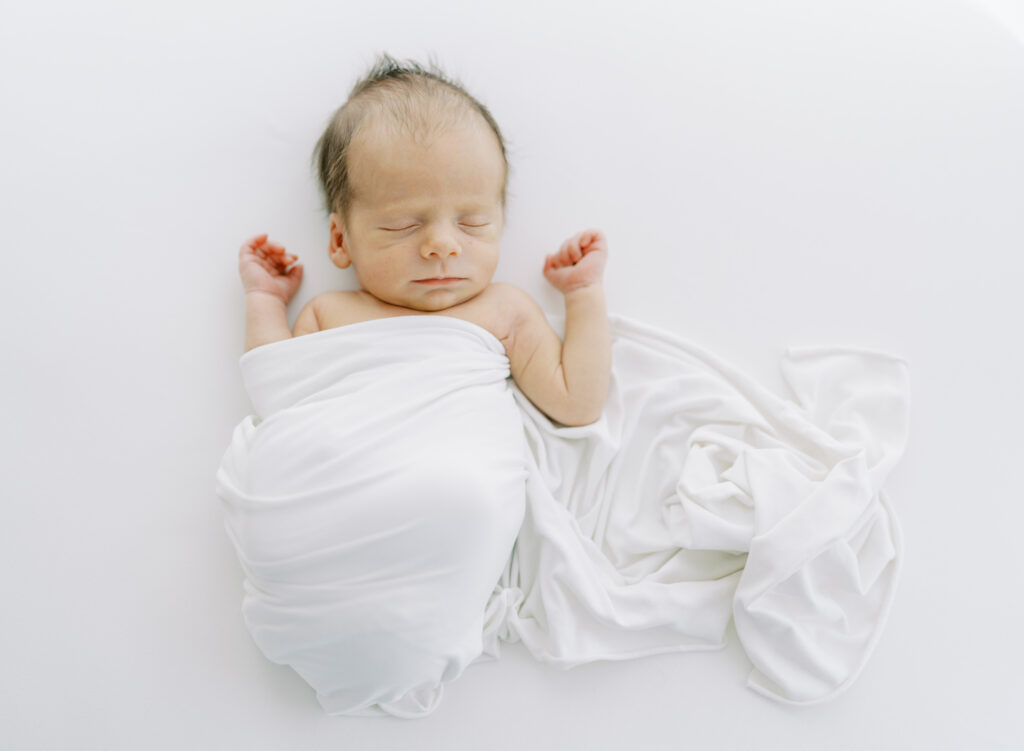 Newborn baby wrapped in a white cloth sleeping on a white bed during newborn photos in Cumming, GA.