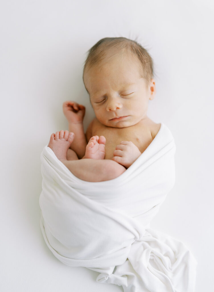 Newborn baby wrapped with his legs up in a white swaddle on a white blanket for a clean and natural newborn photo.